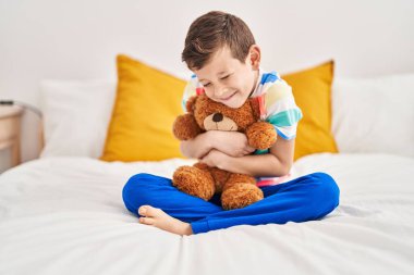 Blond child hugging teddy bear sitting on bed at bedroom
