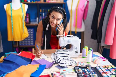 Young beautiful hispanic woman tailor talking on smartphone writing on notebook at clothing factory