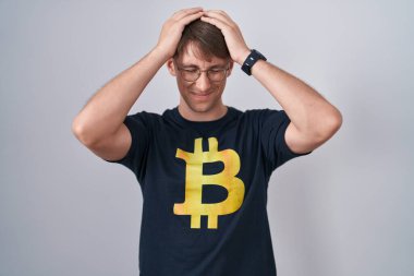 Ağrı ve migren yüzünden baş ağrısı ve stresten muzdarip bitcoin tişörtü giyen beyaz sarışın bir adam. Eller başının üstünde.. 