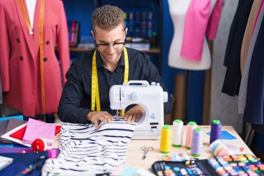 Young caucasian man tailor smiling confident using sewing machine at clothing factory