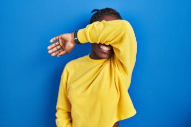 Güzel siyah kadın mavi arka planda dikiliyor. Gözleri kollarıyla kaplıyor. Neşeli ve komik gülümsüyor. kör kavram. 