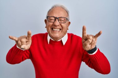 İzole edilmiş arka planda gri saçlı yaşlı adam çılgın bir ifadeyle el kaldırarak rock sembolü yapıyor. Müzik yıldızı. ağır müzik konsepti. 