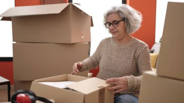 Gri saçlı orta yaşlı bir kadın yeni evinde karton kutuları açıyor.