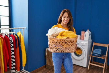 Young latin woman smiling confident holding basket with clothes at laundry room
