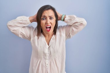 Orta yaşlı İspanyol kadın mavi arka planda çılgın ve korkmuş, elleri başının üstünde, korkmuş ve ağzı açık şoktan şaşırmış bir halde duruyor. 