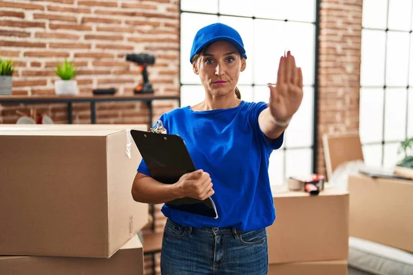 Middle age brunette woman wearing delivery uniform at house moving with open hand doing stop sign with serious and confident expression, defense gesture