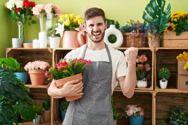 Hispanic man with beard working at florist shop holding plant screaming proud, celebrating victory and success very excited with raised arm