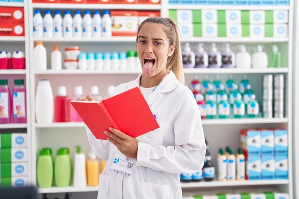 Mulher Loira Jovem Trabalhando Farmácia Farmácia Segurando Notebook Furando Língua — Fotografia de Stock