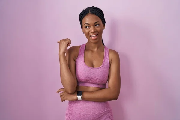 アフリカ系アメリカ人女性とともにスポーツウェアを身に着けていますピンクの背景に笑みを浮かべて幸せな顔を見て 親指で側面を指しています — ストック写真