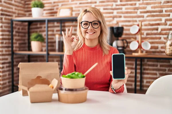ブロンドの女性が食べることは指でOkサインを行うスマートフォンの画面を示す食品を奪う 笑顔優しいジェスチャー優れたシンボル — ストック写真