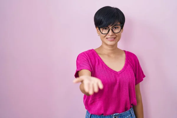 Young asian woman with short hair standing over pink background smiling cheerful offering palm hand giving assistance and acceptance.