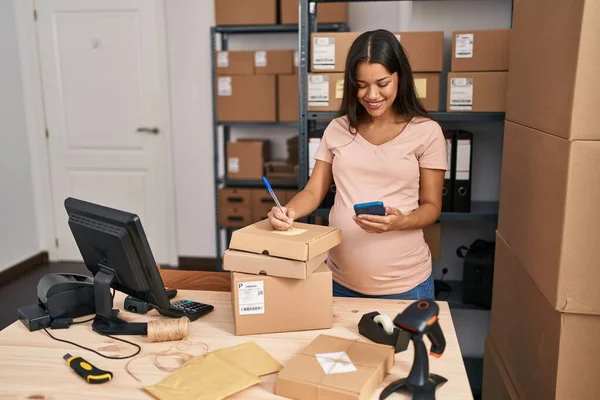 年轻的拉丁裔妇女怀孕了 她在办公室用智能手机在包裹上写字 — 图库照片