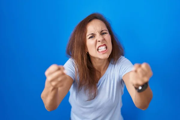 怒りで叫んでいる間に青の背景に立っているブルネットの女性は怒って怒って拳を上げると激怒しました 怒りと攻撃的な考え方 — ストック写真