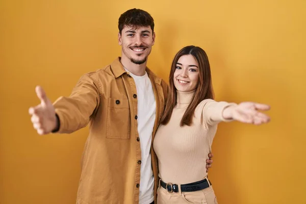 若いヒスパニック系のカップルは カメラを見て黄色の背景に立って抱擁のために腕を開いて笑顔 幸せを受け入れる陽気な表情 — ストック写真
