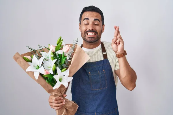 Hispanic man with beard working as florist gesturing finger crossed smiling with hope and eyes closed. luck and superstitious concept.