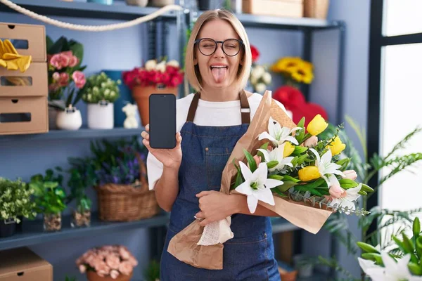 若い白人女性が花屋で働いていますスマートフォンの画面が面白い表情で幸せ舌を突き出しています — ストック写真