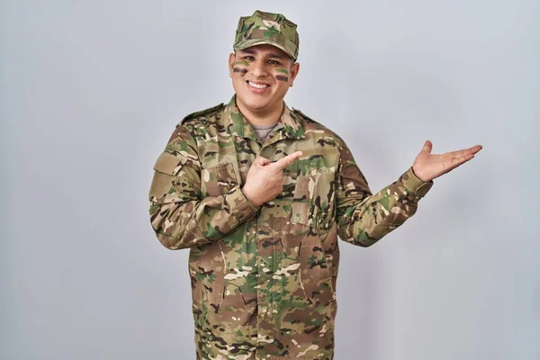 手で提示しながら カメラに驚いて笑顔と指で指差すカモフラージュ軍の制服を身に着けているヒスパニック系の若者 — ストック写真