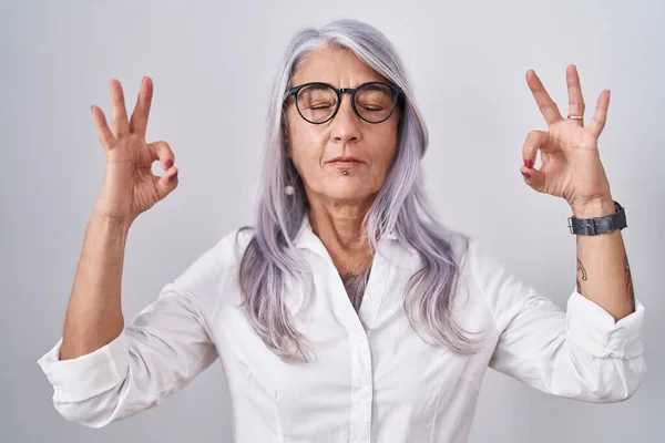 白い背景の上に眼鏡をかけてタトゥーを入れた中年女性がリラックスし 指で瞑想ジェスチャーを行う目で笑顔を閉じます ヨガのコンセプト — ストック写真