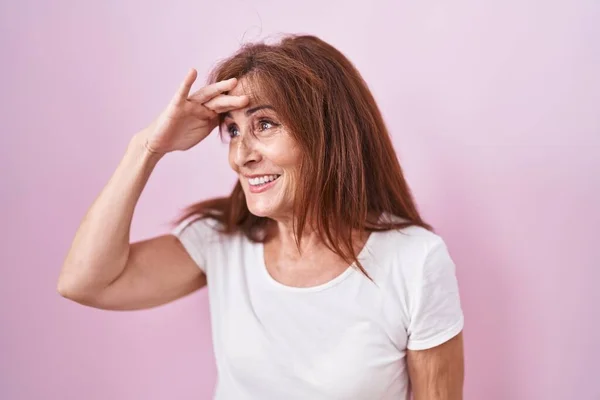 ピンクの背景の上に立っている中年の女性は非常に幸せと遠くに手を頭の上に見て笑顔 検索の概念 — ストック写真