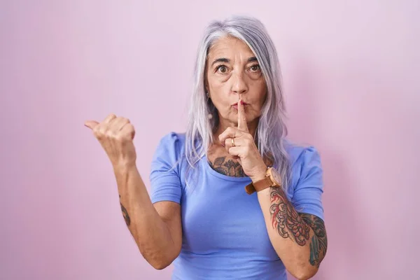 Middle age woman with tattoos standing over pink background asking to be quiet with finger on lips pointing with hand to the side. silence and secret concept.