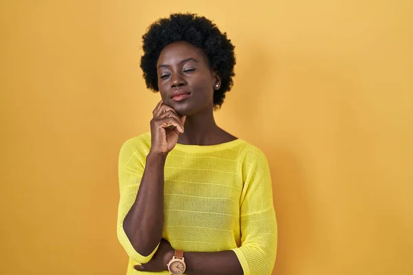 非洲的年轻女子手托着下巴站在黄色的工作室前思考着问题 沉思着自己的表情 脸上挂着沉思的笑容 怀疑概念 — 图库照片