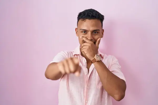 若いヒスパニック系の男がピンクの背景に立ってあなたを笑って 手で口の上にカメラに指を指して 恥の表情 — ストック写真