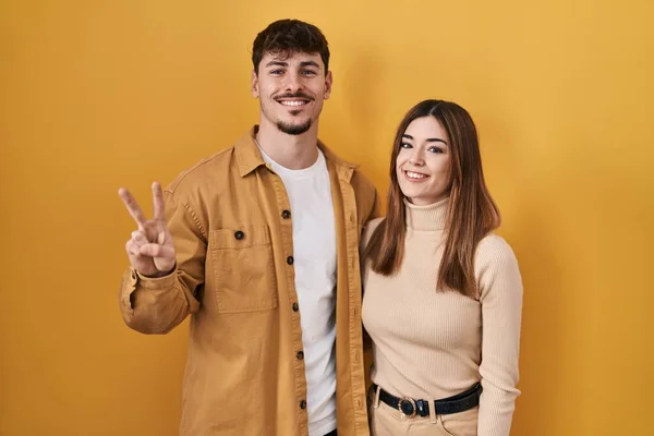 Young hispanic couple standing over yellow background showing and pointing up with fingers number two while smiling confident and happy.