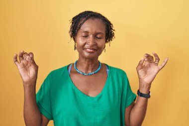 Sarı arka planda dikilen Afrikalı kadın rahatlamış ve parmaklarıyla meditasyon hareketi yaparken gözleri kapalı gülümsüyor. yoga konsepti. 