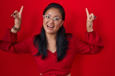 Asyalı genç bir kadın kırmızı arka planda duruyor. Gülümsüyor, şaşırıyor, parmaklarıyla işaret ediyor ve kollarını kaldırıyor.. 