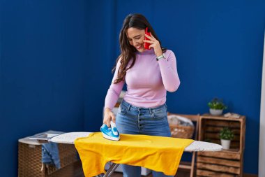 Young hispanic woman talking on the smartphone ironing clothes at laundry room
