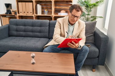 Middle age man writing on clipboard having psychology session at clinic