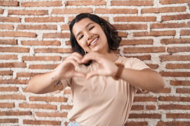 Tuğla duvarın yanında dikilen genç İspanyol kadın. Sevgiyle gülümsüyor. Elleriyle kalp sembolü yapıyor. romantik kavram. 