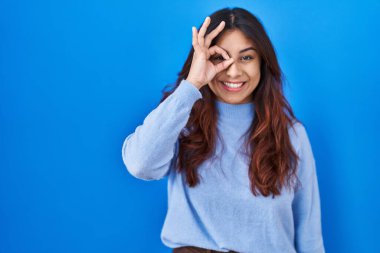 Latin kökenli genç bir kadın mavi arka planda duruyor. El hareketleriyle gülümsüyor, gözleriyle mutlu yüzlere bakıyor.. 