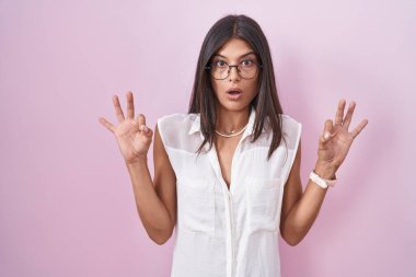 Pembe arka planda duran esmer, gözlüklü, şaşırmış ve şok olmuş genç bir kadın parmaklarıyla onay sembolü yapıyor. çılgın ifade 