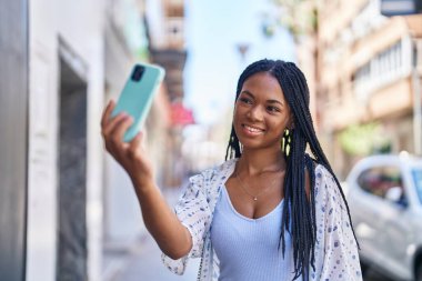 Afro-Amerikalı kadın gülümsüyor. Sokakta akıllı telefonun yanında özçekim yapıyor.