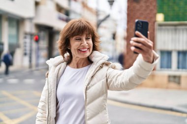 Orta yaşlı kadın gülümsüyor. Sokakta akıllı telefonun yanında özçekim yapıyor.