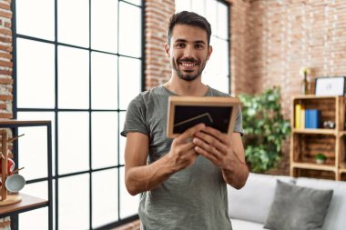 Young hispanic man smiling confident looking picture frame at home