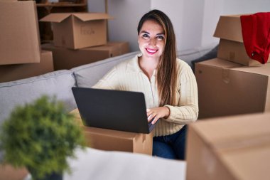 Young beautiful hispanic woman using laptop sitting on sofa at new home