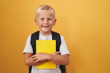 Küçük beyaz çocuk öğrenci çantası takıyor ve elinde kitap tutuyor. Pozitif ve mutlu görünüyor. Dişlerini gösteren bir gülümsemeyle ayakta duruyor ve gülümsüyor. 