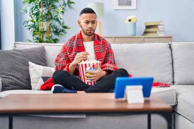 Genç İspanyol bir adam tablet cihazında patlamış mısır yiyor şüpheci ve gergin, sorun yüzünden surat asıyor. negatif kişi. 