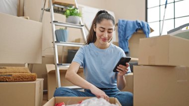 Genç, güzel, İspanyol bir kadın yeni evinde akıllı telefon kullanarak karton kutuları açıyor.