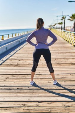 Young beautiful hispanic woman wearing sportswear standing on back view at seaside