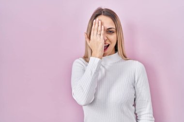 Pembe arka planda beyaz süveter giyen genç sarışın kadın bir gözünü eliyle kapatıyor, yüzünde kendine güvenen bir gülümseme ve sürpriz bir duygu var.. 