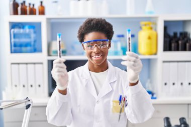 Laboratuvarda test tüpleri tutan bilim adamı üniforması giyen Afrikalı Amerikalı bir kadın.