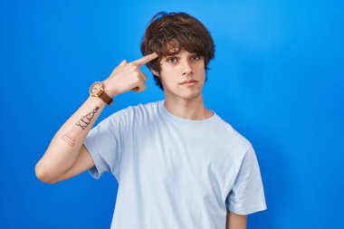 Mavi arka planda duran İspanyol genç adam alnındaki sivilceden mutsuz olduğunu gösteriyor, siyah başlıklı çirkin bir enfeksiyon. sivilce ve deri problemi 