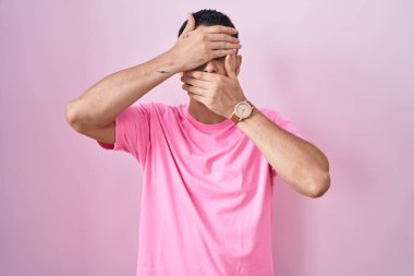 Pembe arka planda duran İspanyol genç adam gözlerini ve ağzını ellerle kapadı, şaşırdı ve şok oldu. duygu gizleme 