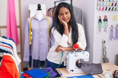 Young beautiful woman tailor smiling confident leaning on sewing machine at tailor shop