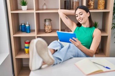 Young hispanic woman using touchpad sitting on table at home
