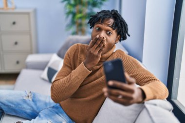 Rastalı saçlı genç Afrikalı bir adam akıllı telefon kullanarak ağzını eliyle kapatıyor, şok geçiriyor ve hata yapmaktan korkuyor. şaşırmış ifade 