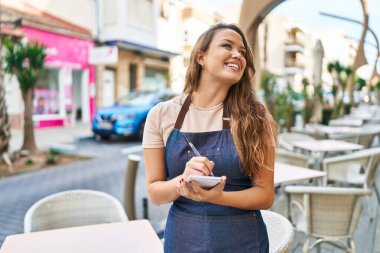 Young beautiful hispanic woman waitress smiling confident writing on notebook at coffee shop terrace